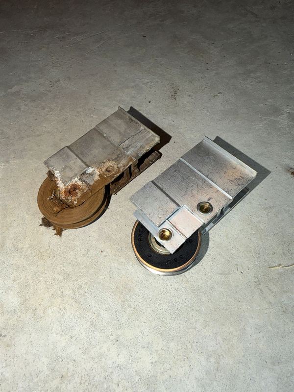 Old rusted sliding door roller next to a brand new stainless steel roller assembly on a concrete slab