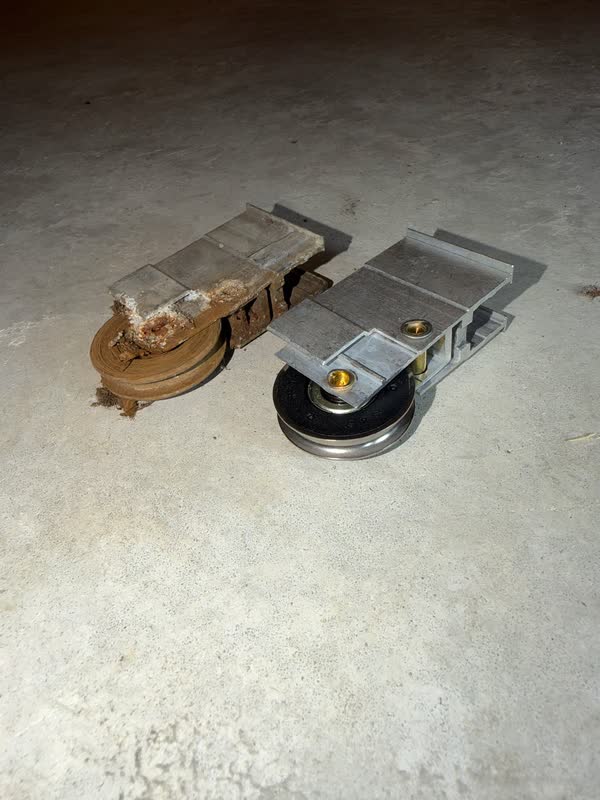 A rusted, seized old sliding door roller next to a brand-new sealed stainless steel replacement roller on a concrete floor